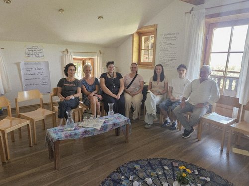 Sieben Personen sitzen in einem hellen Raum mit Holzboden nebeneinander auf Stühlen. Im Hintergrund hängen Tafeln mit Notizen. In der Mitte steht ein niedriger Tisch mit einer Karaffe und Gläsern.