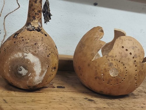 Zwei getrocknete Kalebassen auf einem Holzbrett. Links steht ein ganzer Flaschenkürbis mit langem Hals und gesprenkelter Schale. Rechts daneben liegt eine runde, ausgehöhlte Kalebasse, die dekorativ mit kleinen Löchern in Spiralmustern durchbohrt ist.