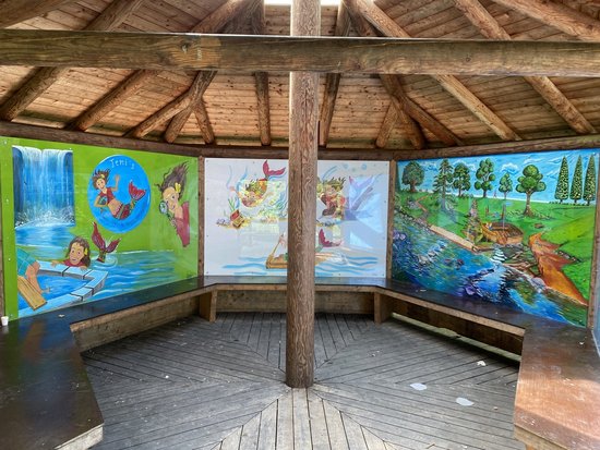 Innenansicht eines offenen Holz-Pavillons mit farbenfrohen Wandgemälden. Die Kunstwerke stellen Natur- und Wasserszenen dar, einschließlich Wasserfällen, schwimmenden Kindern und Personen, die auf einem Fluss flößen. Der Raum hat einen Holzboden, ein Dach aus Holzbalken und lange Bänke entlang der Wände.