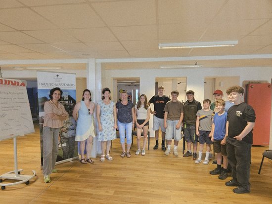 Eine Gruppe von etwa elf Erwachsenen und Jugendlichen steht in einer Reihe in einem Raum mit Holzboden und posiert für ein Foto. Im Hintergrund sind zwei Banner zu sehen, eines davon mit der Aufschrift „Haus Schwarzenburg“.