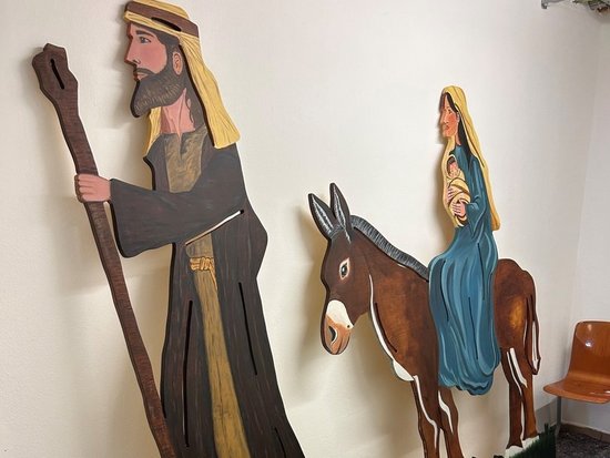 Wandmontierte Holzfiguren stellen Josef und Maria dar. Josef steht links mit einem Wanderstab, während Maria rechts auf einem braunen Esel reitet. Die flachen Figuren sind an einer weißen Wand befestigt und stehen auf einer grünen Grasbasis.