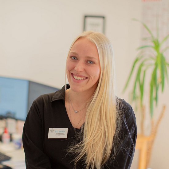 Eine junge Frau mit langem blonden Haar sitzt locker auf einem Schreibtisch im Büro und lächelt in die Kamera