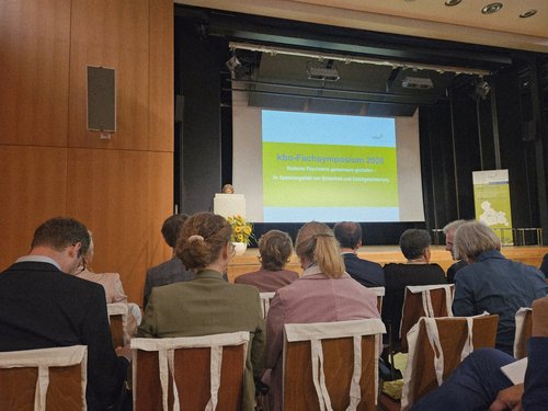 Ein Publikum sitzt in einem Saal und blickt auf eine Bühne. Dort steht ein Redner am Podium, und auf einer großen Leinwand ist eine Präsentation mit dem Titel „Fachtagung 2018" zu sehen.