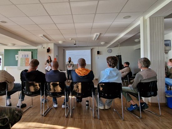 Eine Gruppe von Menschen sitzt in einem Raum mit Holzboden und blickt auf eine Projektionsfläche, wo zwei Personen stehen und einen Vortrag halten. Im Vordergrund ist eine Person in einem orangen Hoodie zu sehen.