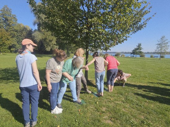 Eine Gruppe von Menschen steht auf einer Wiese unter einem Baum und bildet eine Kette, indem sie sich an den Händen hält. Im Hintergrund ist ein See bei sonnigem Wetter zu sehen.