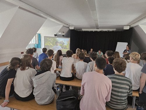 Jugendliche sitzen in einem Seminarraum mit schräger Decke auf Bänken und blicken auf eine Projektionsfläche. Ein Referent steht rechts und hält einen Vortrag, während auf der Leinwand eine Naturlandschaft gezeigt wird.