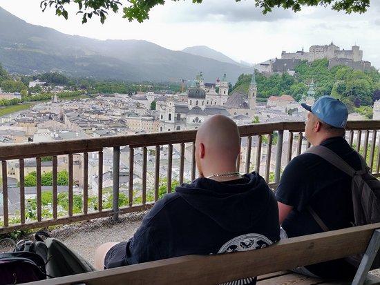 Zwei Männer sitzen auf einer Holzbank und blicken über die historische Altstadt von Salzburg, den Fluss und die Festung Hohensalzburg auf dem Berg.