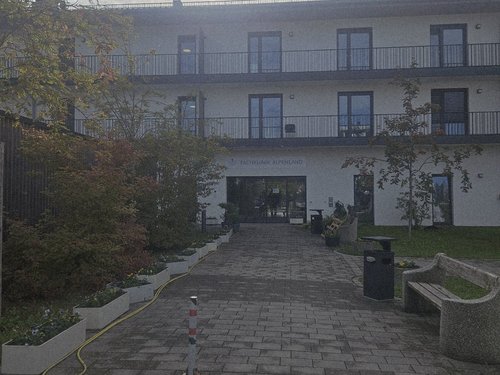 Das Bild zeigt die Außenansicht eines weißen, mehrstöckigen Gebäudes mit Balkonen, vermutlich ein Hotel. Ein gepflasterter Weg führt zum Eingang. Bäume säumen den Weg, und die Sonne scheint hell am bewölkten Himmel.