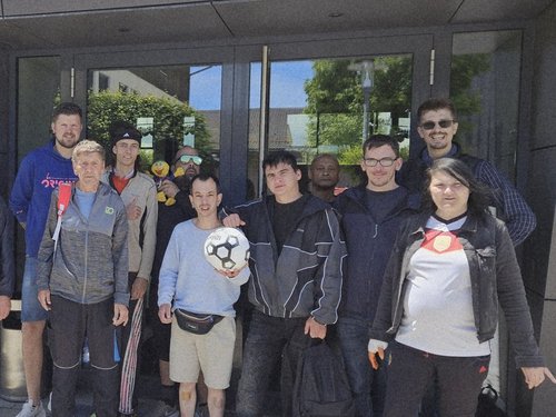 Eine Gruppe von etwa zehn Personen steht lächelnd vor dem Eingang eines modernen Gebäudes mit Holz- und Graufassade. Eine Person in der Mitte hält einen Fußball. Links im Bild ist ein Fahrrad abgestellt.