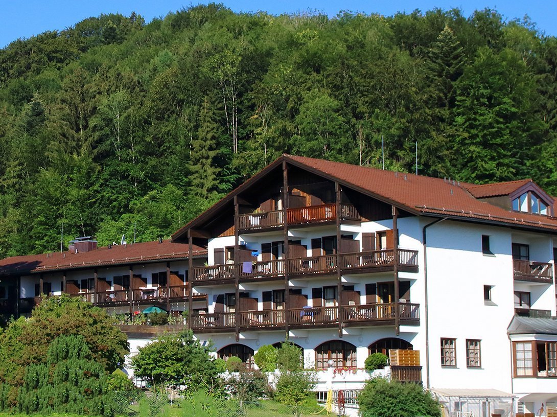 Aussenansicht: ein großes Gebäude mit vielen Balkonen und einer großen Terasse umringt von vielen Bäumen an einem leichten Hang