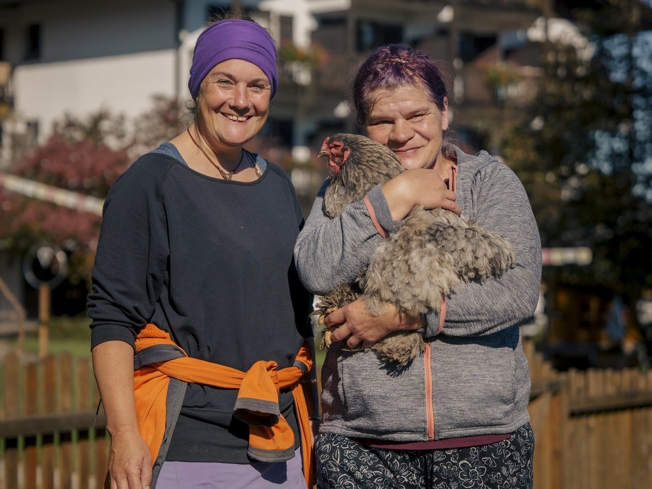 zwei Frauen lachen in die Kamera währenddessen sie ein Huhn auf dem Arm haben 