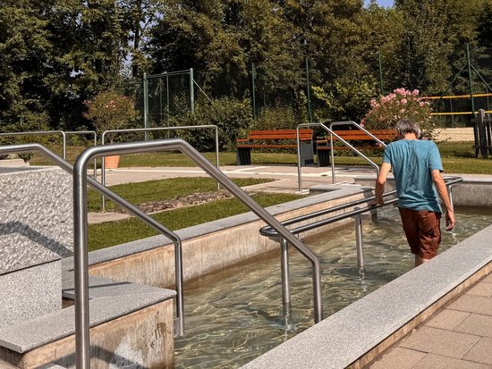 Ein Mann in blauem T-Shirt und braunen Hosen geht barfuß in ein flaches Wasserbecken mit Metallgeländern, vermutlich eine Kneipp-Anlage im Park. Im Hintergrund sind grüne Bäume und Parkbänke unter einem klaren blauen Himmel zu sehen.