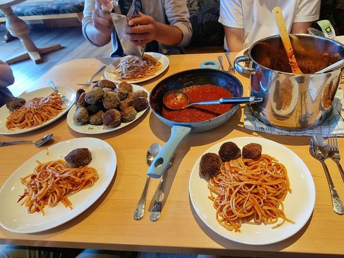Mehrere Teller mit Spaghetti und Frikadellen stehen auf einem Holztisch. In der Mitte befinden sich eine große Pfanne mit Tomatensauce und ein Kochtopf. Personen sitzen um den Tisch herum und essen gemeinsam.