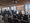 In einem hellen Raum mit Panoramafenstern steht eine Gruppe von Menschen vor einer Projektionsleinwand mit der Aufschrift „20 Jahre“. Im Vordergrund sind Reihen leerer schwarzer Stühle angeordnet, was auf eine Jubiläumsveranstaltung oder Konferenz hindeutet.