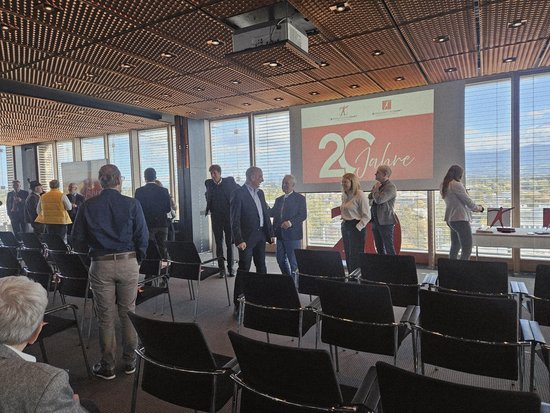 In einem hellen Raum mit Panoramafenstern steht eine Gruppe von Menschen vor einer Projektionsleinwand mit der Aufschrift „20 Jahre“. Im Vordergrund sind Reihen leerer schwarzer Stühle angeordnet, was auf eine Jubiläumsveranstaltung oder Konferenz hindeutet.