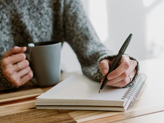 Eine Person in einem grauen Strickpullover sitzt an einem Holztisch und schreibt mit einem Stift in ein Notizbuch, während sie eine Tasse in der anderen Hand hält.
