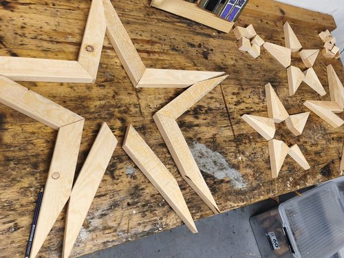 Auf einer Holzwerkbank sind verschiedene hölzerne Sternformen zu sehen. Neben großen, flachen Sternrahmen liegen mehrere kleinere, dreidimensionale geometrische Sterne, die aus einzelnen Holzstücken zusammengesetzt sind.